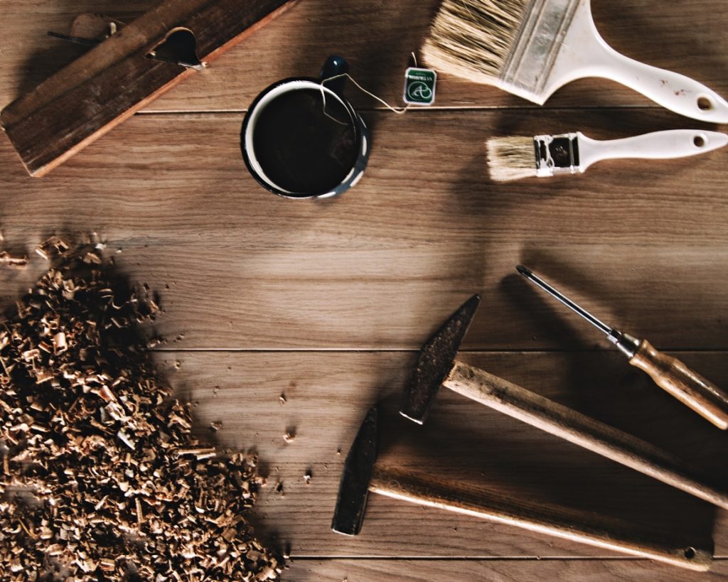 Drewniany blat a na nim narzędzia: dwa młotki, śrubokręt, pędzle oraz trociny i kubek z herbatą.