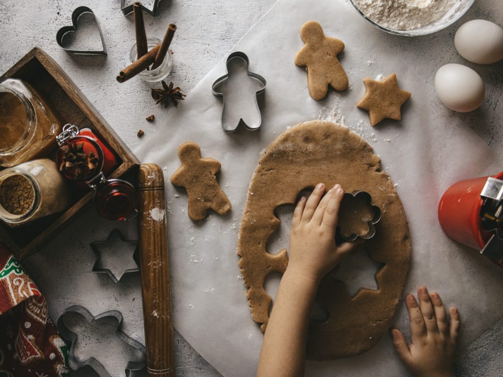 Ręce dziecka wycinające z płatu ciasta pierniczki o różnych kształtach; składniki na ciasto na pierniczki: jajka, mąka, kakao, cynamon, gwiazdki anyżu.
