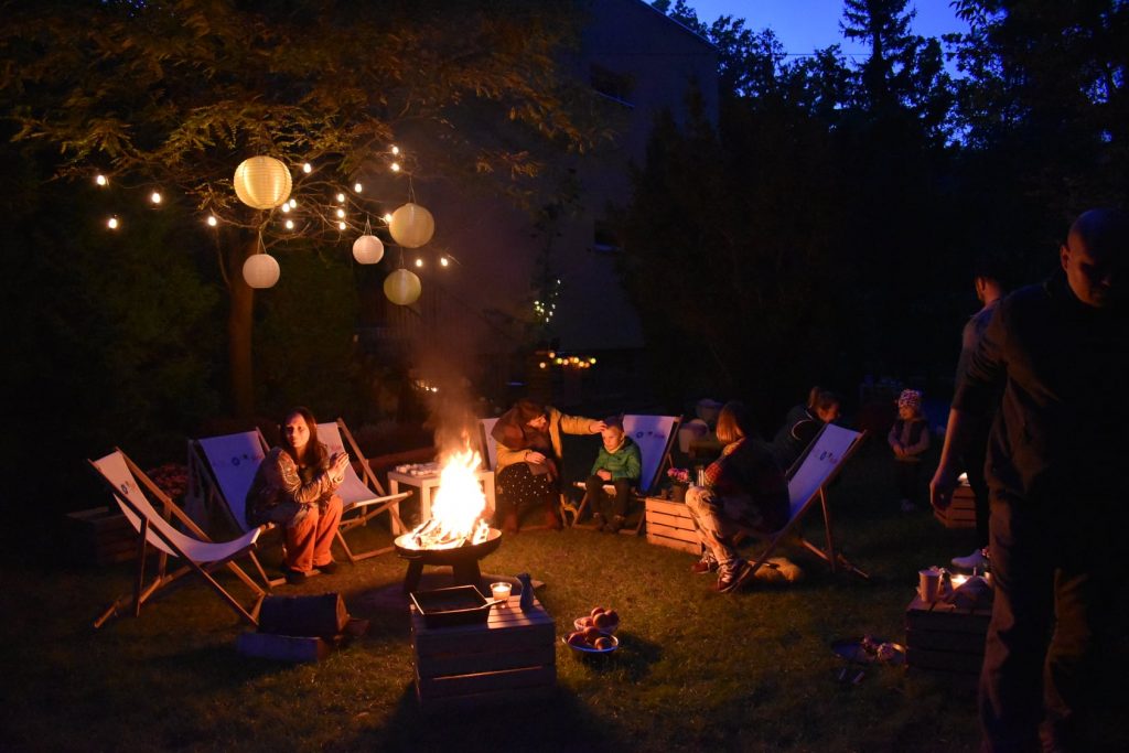 "Dzień Ziemniaka" w ogrodzie MAL Samogłoska: ludzie siedzący na leżakach wokół ogniska, lampiony i girlanda świetlna rozwieszone na drzewie.