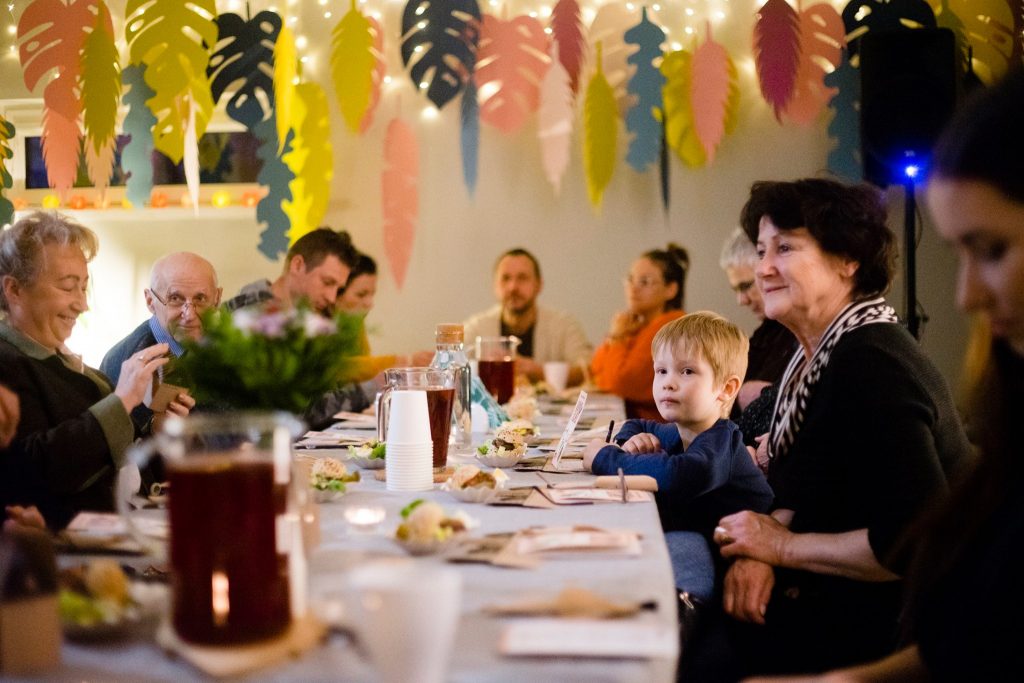 Kobiety, mężczyźni i dziecko siedzący przy stole w MAL Samogłoska podczas wydarzenia "Korzenie Młocin".