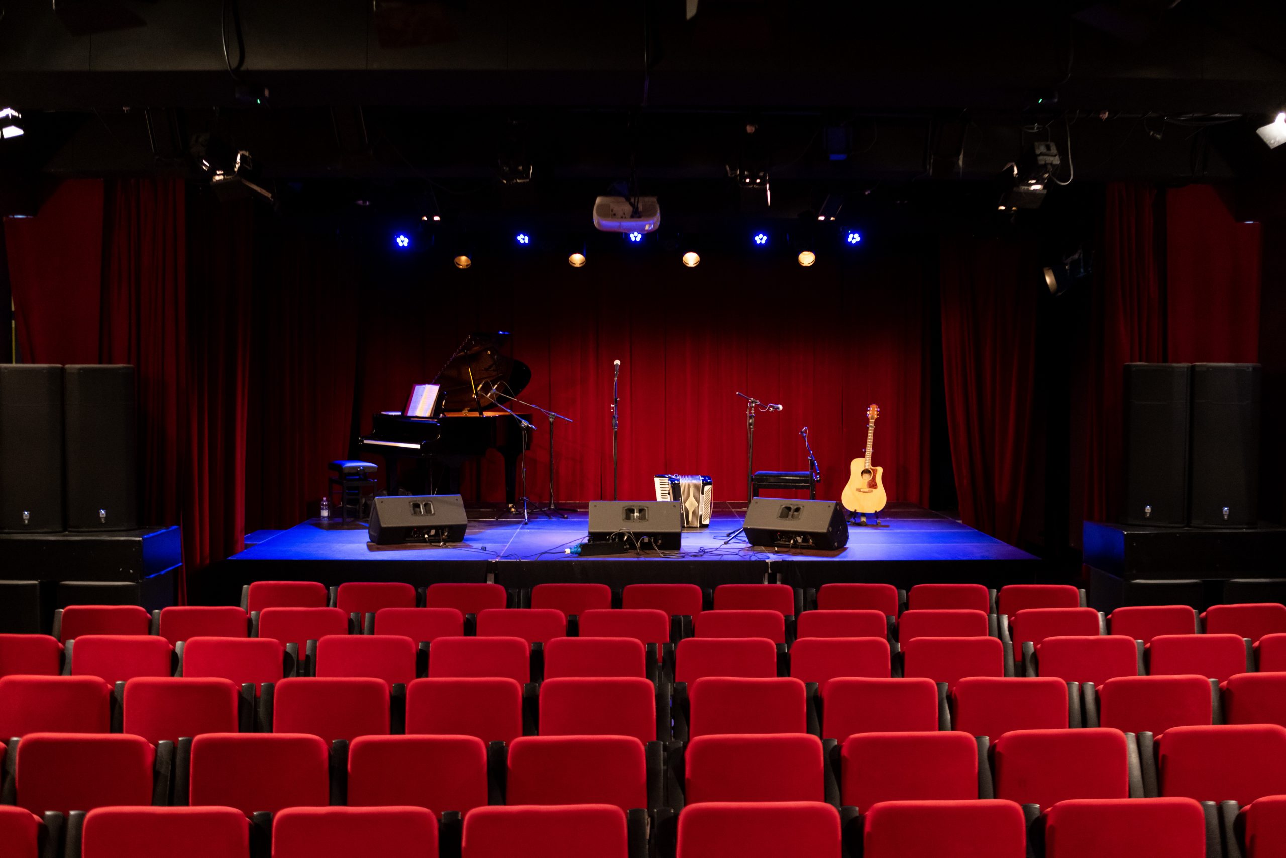 Sala widowiskowa Bielańskiego Ośrodka Kultury przy ulicy Goldoniego 1. Rzędy bordowych krzeseł i scena przygotowana do koncertu.