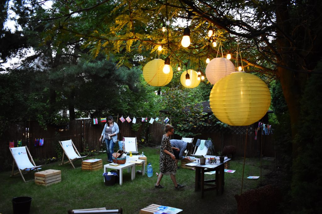 Warsztaty marblingu w ogrodzie MAL Samogłoska; leżaki i drewniane skrzynki rozstawione na trawie, lampiony solarne i girlanda świetlna rozwieszone na drzewie.
