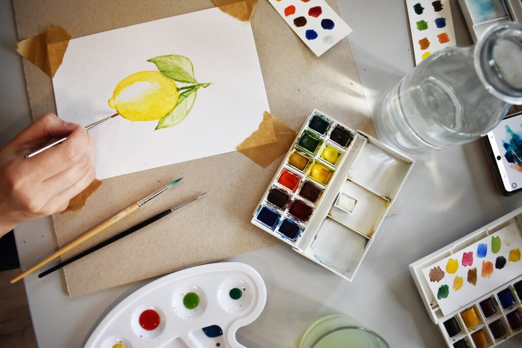 Akwarelowy obrazek żółtej cytryny, palety z akwarelami i paletki do mieszania farb.