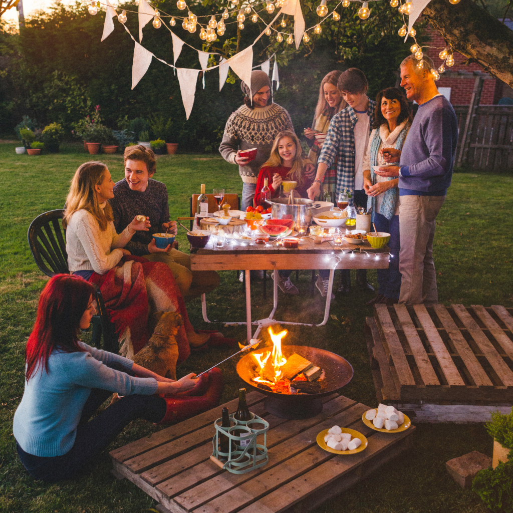 Roześmiani ludzie zgromadzeni wokół stołu z jedzieniem w ogrodzie, kobieta piekąca piankę na ognisku.