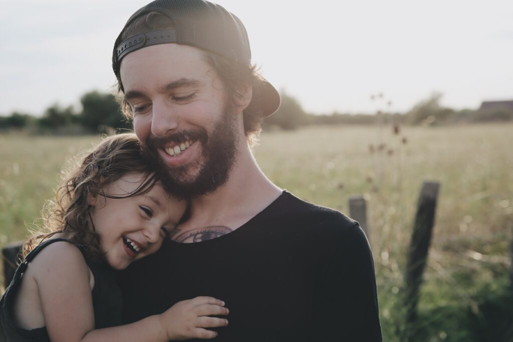Mężczyzna i dziewczynka przytuleni do siebie i uśmiechający się na tle łąki.