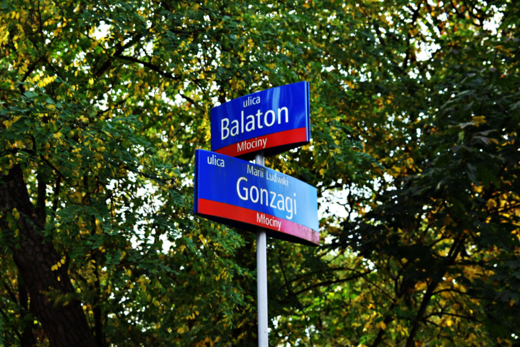 Znak z nazwami ulic na Młocinach: ulicą Balaton i Gonzagi na tle zielonych drzew.