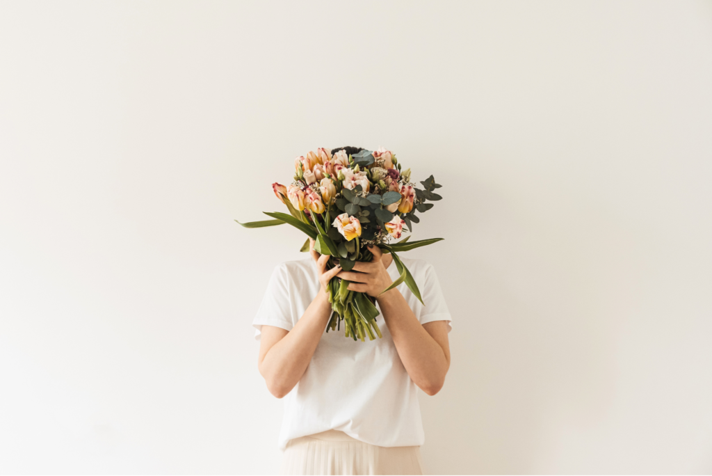 Kobieta na tle jasnej ściany trzyma bukiet kwiatów na wysokości swojej twarzy.