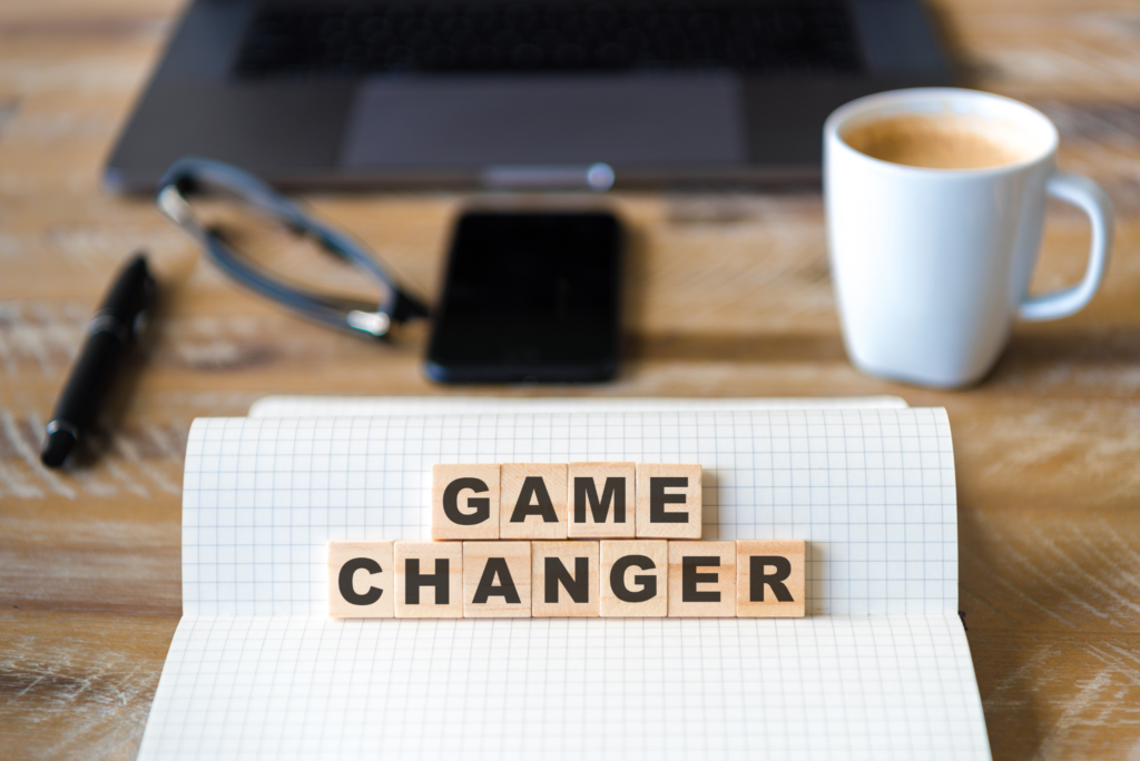 Obrazek przedstawia napis z drewnianych klocków 'Game Changer" postawiony na otwartym zeszycie w kratkę, w tle kubek, telefon, okulary, laptop i długopis
