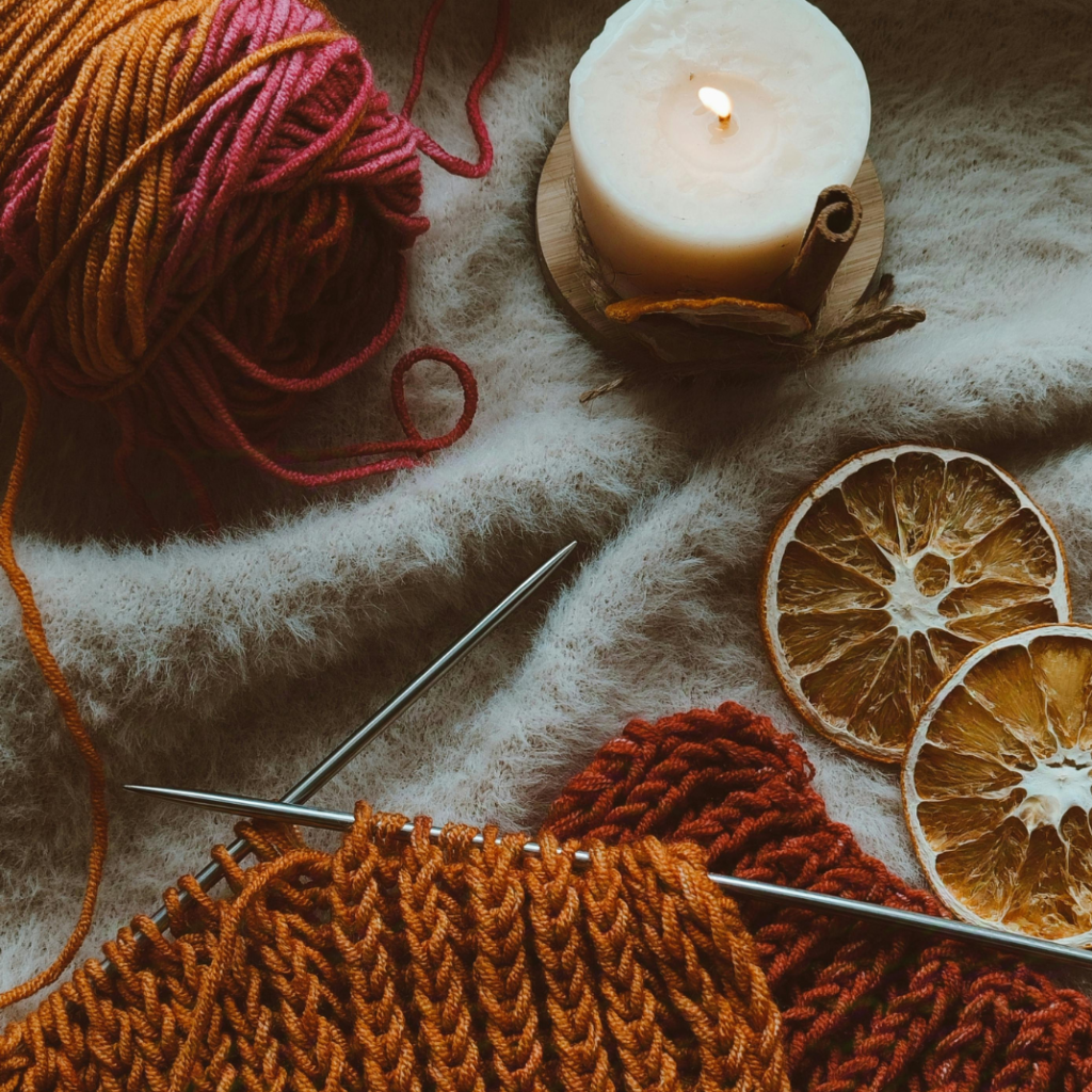 Pomarańczowa i czerwona włóczka, metalowe druty, świeczka i ususzone pomarańcze na tle beżowego koca.
